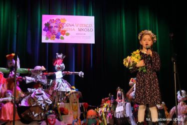 Na scenie dziewczynka z wiankiem na głowie, w prawej ręce trzyma bukiet żółtych kwiatów a w  lewej mikrofon recytuje wiersz.