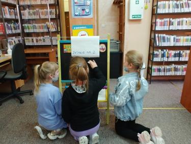 Trzy dziewczynki klęczą przy tablicy do pisania, jedna z nich trzyma w ręce kredę 