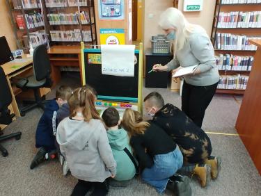 Uczniowie klęczą na podłodze i patrzą na tablicę do rozwiązywania zagadki, obok stoi ich nauczycielka z zeszytem i długopisem w ręce