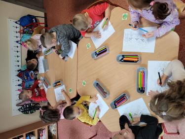  Przy dwóch złączonych stolikach siedzą dzieci przedszkolne, malują kredkami wydrukowane kartki kolorowanki, obok kolorowanek stoją plastikowe pojemniczki z kredkami.