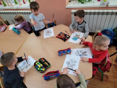 Dzieci przy stoliku kolorują wydrukowane kształty piernika.