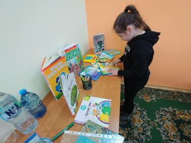Na biurku leżą książeczki dla dzieci, obok stoją dwie butelki z wodą, przy biurku stoi dziewczynka która koloruje zieloną kredką wydrukowany z papieru listek.